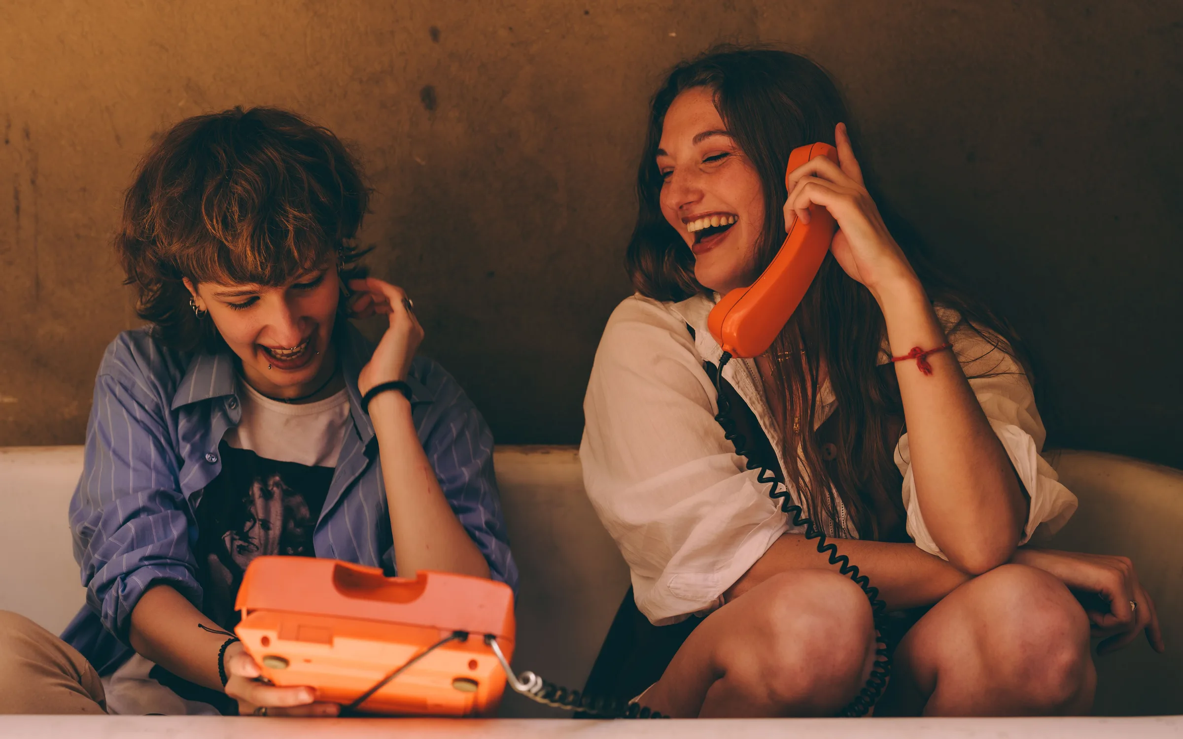 Two friends laughing together, one holding the orange handset of an old telephone, the other its base on her lap.